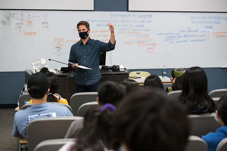 A professor teaching a class with a whiteboard in the background.