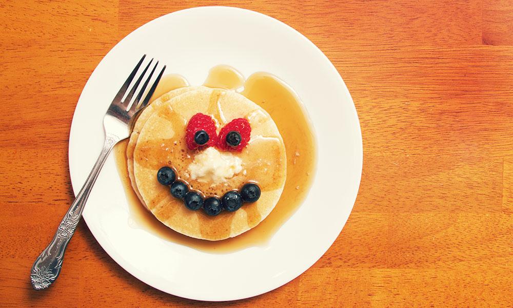 a plate of pancakes and maple syrup making a funny face with fruit..