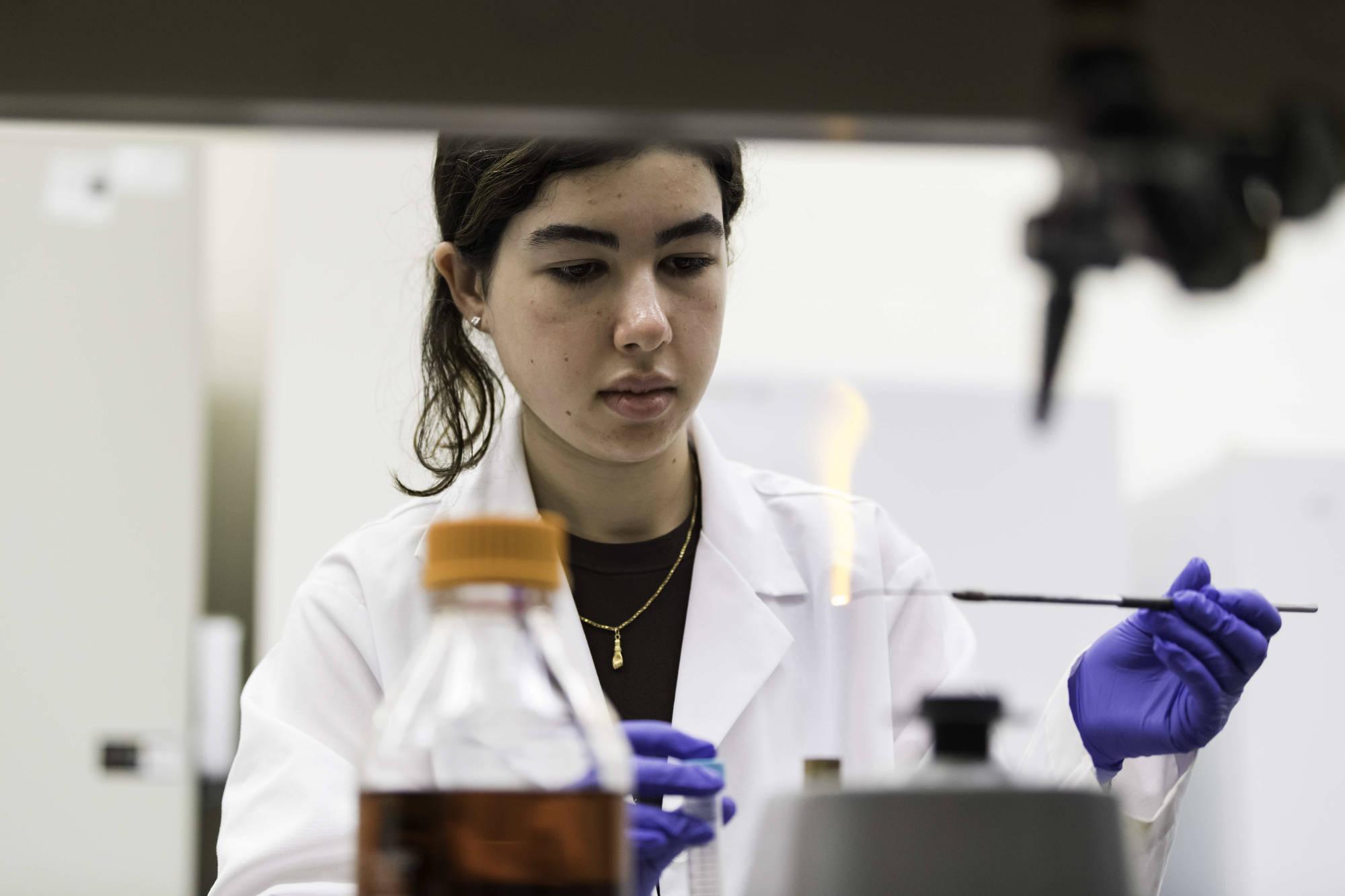 Student in a lab coat sterilizing an inoculation loop using a flame.