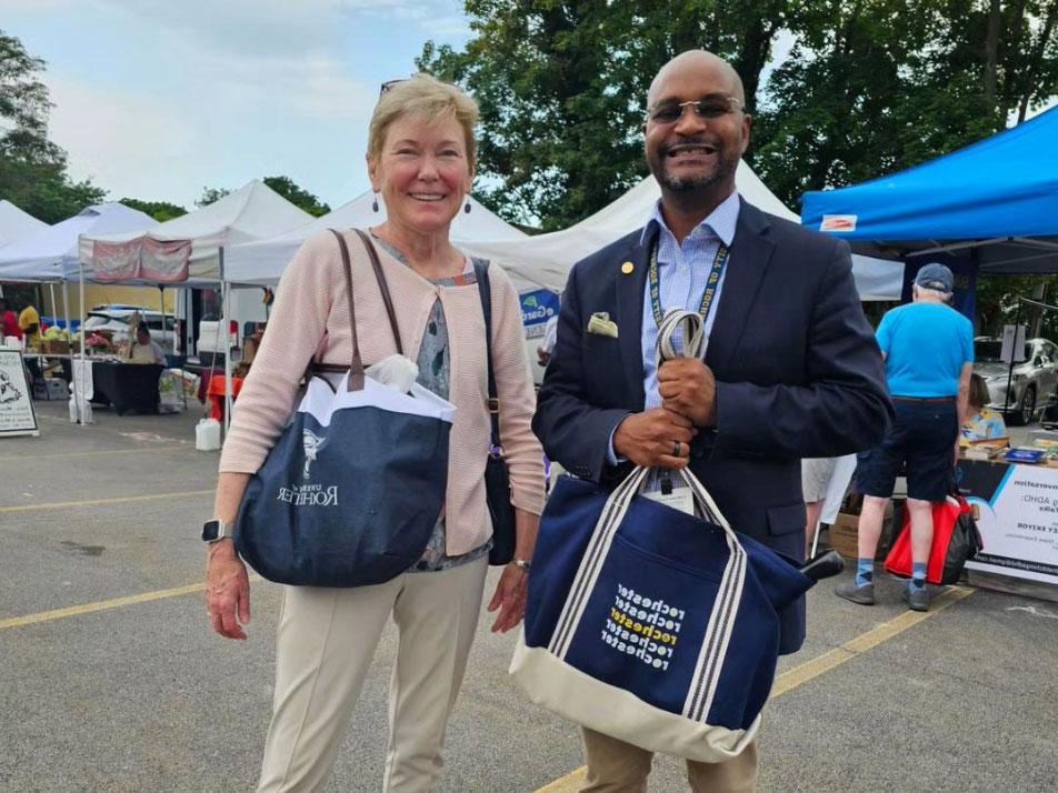 Two people smiling at a market with tents in the background, holding tote bags from the University of Rochester.